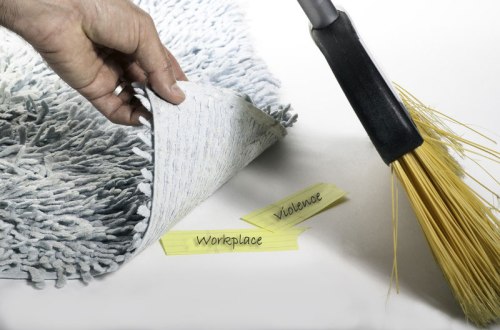 Photograph of person sweeping two post-it notes under a rug. One says Workplace, the other violence.